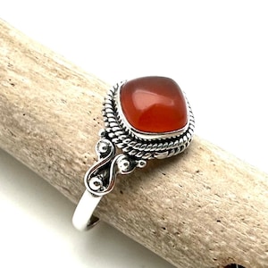May include: A silver ring with a square, reddish-orange carnelian gemstone. The stone is set in a detailed, rope-like bezel. The ring's band has decorative flourishes on the sides. The ring is displayed on a piece of driftwood.