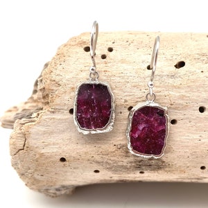 May include: A pair of silver earrings with a red ruby stone in each. The earrings are hanging from a piece of driftwood.