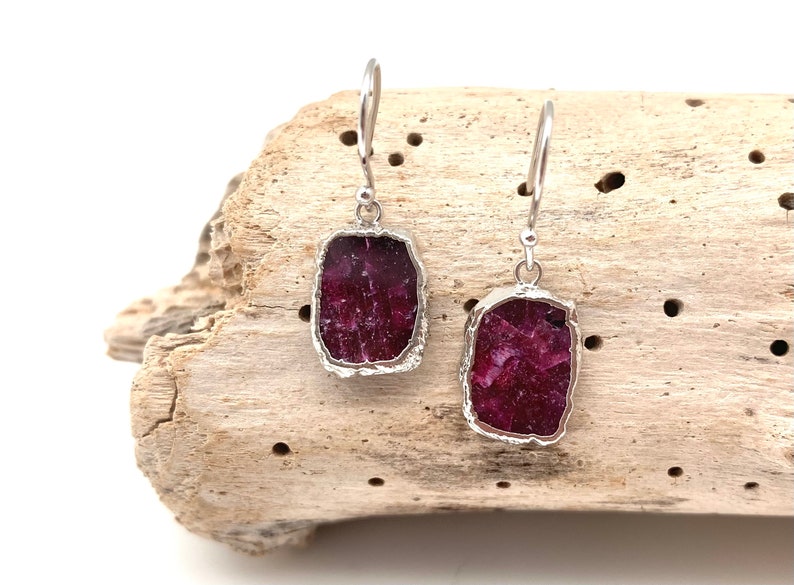 May include: A pair of silver earrings with a red ruby stone in each. The earrings are hanging from a piece of driftwood.