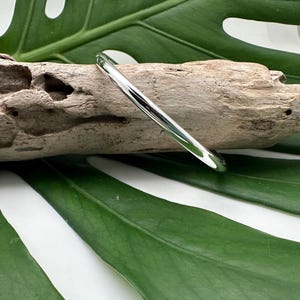 May include: A silver bangle bracelet with a smooth, polished finish. The bracelet is lying on a piece of driftwood and a green leaf.