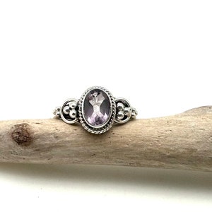 May include: A silver ring with an oval-shaped amethyst gemstone set in a beaded bezel. The ring has a delicate design with small silver beads on the band.
