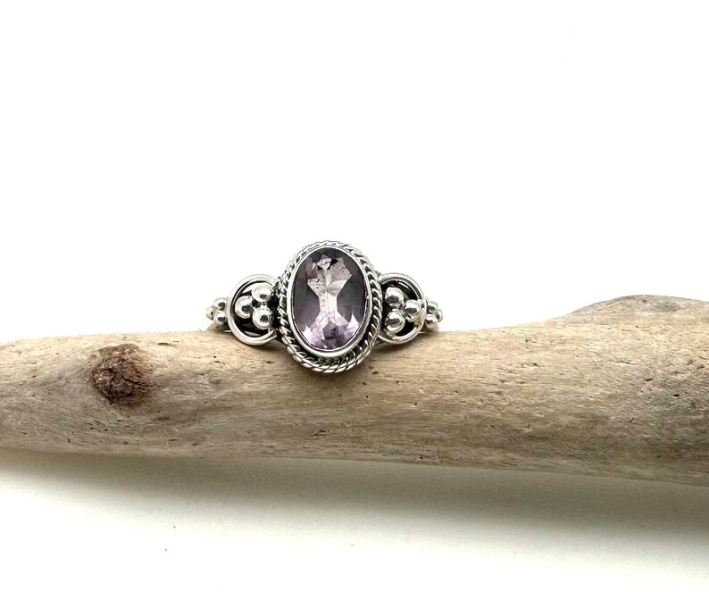 May include: A silver ring with an oval-shaped amethyst gemstone set in a beaded bezel. The ring has a delicate design with small silver beads on the band.