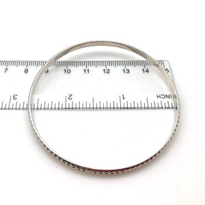 May include: A silver bangle bracelet with a textured design, measured against a ruler. The bracelet is approximately 2.75 inches in diameter.
