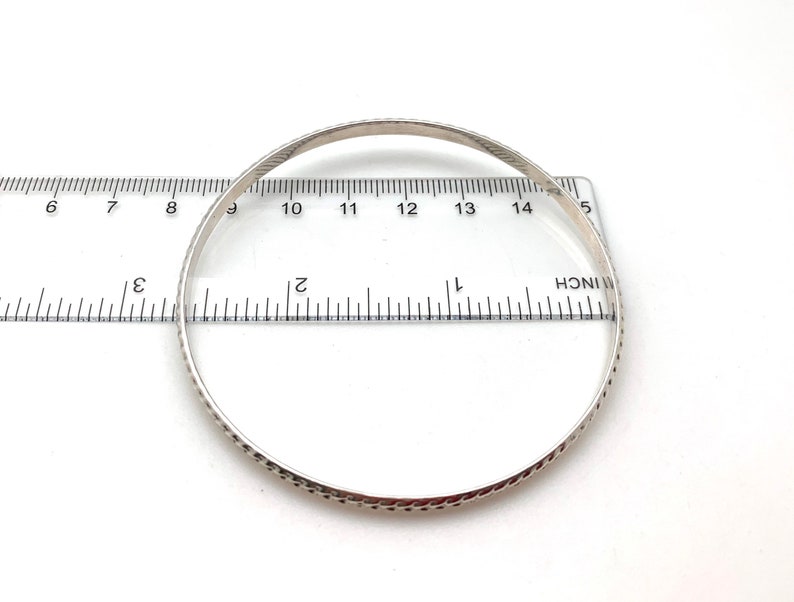 May include: A silver bangle bracelet with a textured design, measured against a ruler. The bracelet is approximately 2.75 inches in diameter.