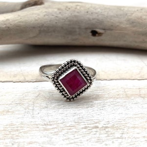 Op de afbeelding: Een zilveren ring met een vierkante robijn-edelsteen gezet in een decoratieve rand.