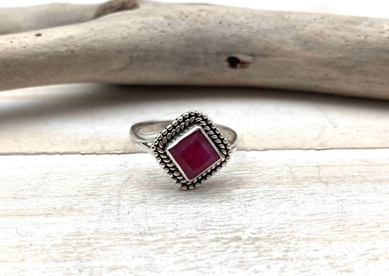 Op de afbeelding: Een zilveren ring met een vierkante robijn-edelsteen gezet in een decoratieve rand.