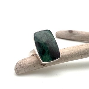 May include: A rectangular ring with a dark green stone set in a silver band. The stone has a mottled appearance with shades of green and black. The ring is displayed on a piece of driftwood against a white background.