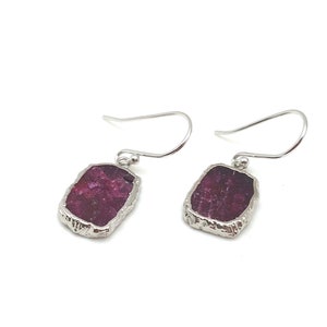 May include: A pair of silver earrings with a raw, rectangular ruby gemstone in each. The ruby is a deep red color and has a rough, natural texture.