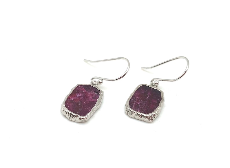 May include: A pair of silver earrings with a raw, rectangular ruby gemstone in each. The ruby is a deep red color and has a rough, natural texture.
