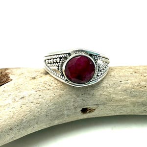 May include: A silver ring with a red gemstone set in a decorative bezel. The ring has a braided design on the band.