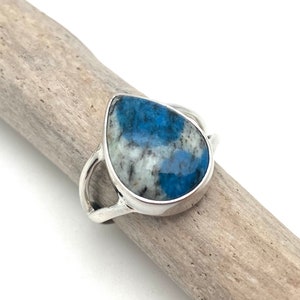 Peut inclure: Une bague en argent avec une pierre pr&eacute;cieuse bleu et blanc en forme de larme. La bague est pos&eacute;e sur un morceau de bois flott&eacute;.