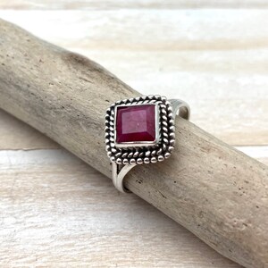 Op de afbeelding: Een zilveren ring met een vierkante robijn-edelsteen gezet in een decoratieve rand. De ring ligt op een houten oppervlak.