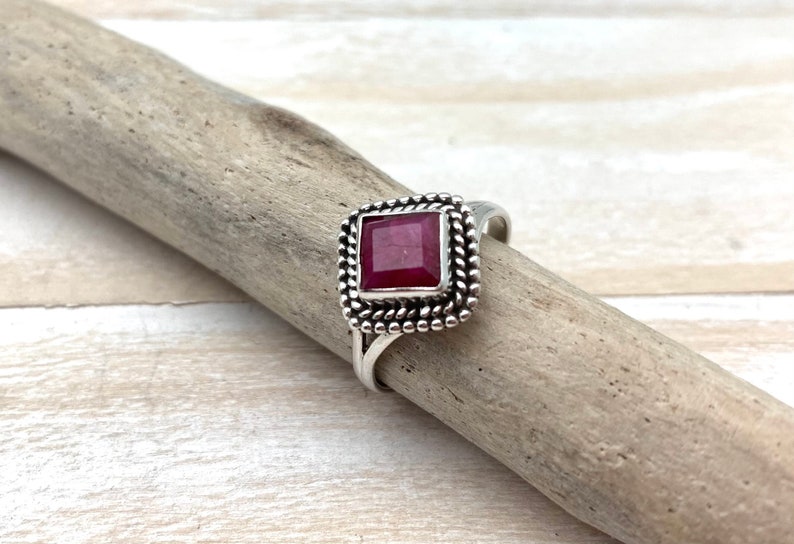 Op de afbeelding: Een zilveren ring met een vierkante robijn-edelsteen gezet in een decoratieve rand. De ring ligt op een houten oppervlak.