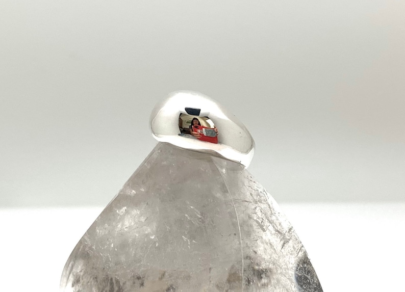 May include: A smooth, silver ring with a rounded, dome-shaped top. The ring is sitting on a white, textured surface.