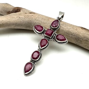 Puede incluir: Un colgante de cruz de plata con piedras preciosas de rubí rojo. La cruz está adornada con siete piedras de rubí, incluida una piedra de forma cuadrada en el centro.