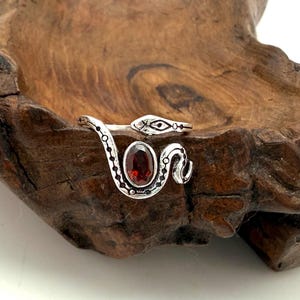 Peut inclure: Une bague argentée en forme de serpent enroulé avec un design ouvert. Le serpent présente des détails noirs gravés le long de son corps et une pierre précieuse rouge foncé de forme ovale sertie au centre de la partie avant de la bague.