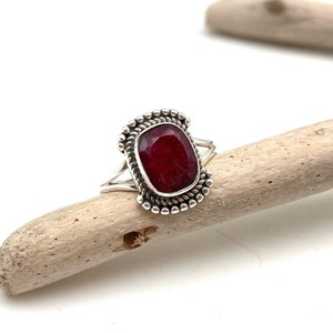 May include: A silver ring with a dark red gemstone set in a decorative silver bezel. The ring is on a piece of driftwood.