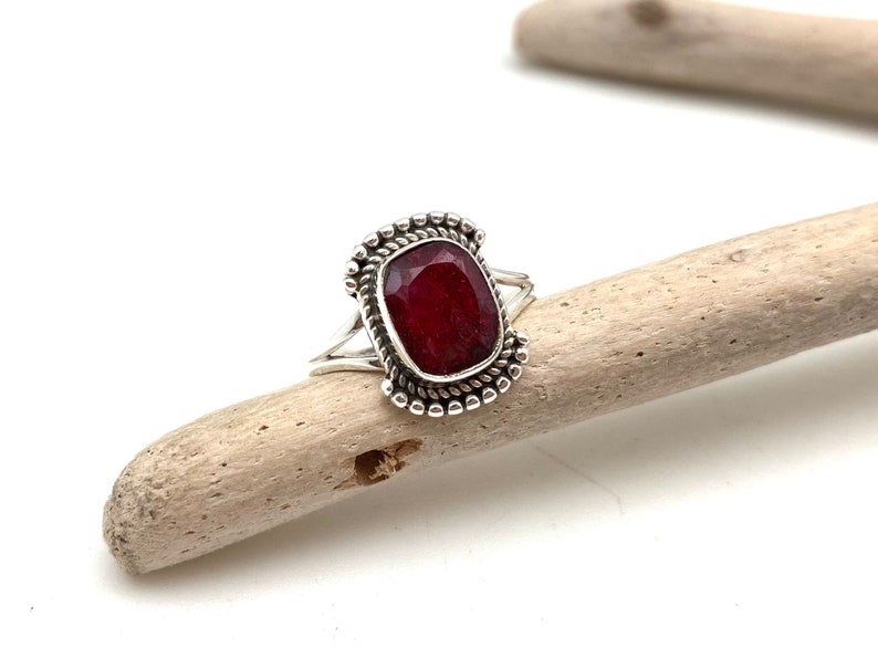 Op de afbeelding: Een zilveren ring met een donkerrode edelsteen gezet in een decoratieve zilveren zetting. De ring ligt op een stuk drijfhout.