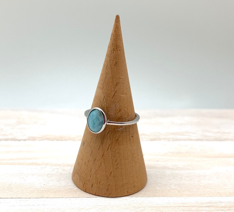 May include: A silver ring featuring a light blue oval gemstone. The ring is displayed on a wooden ring stand.