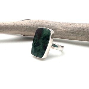 May include: A rectangular ring with a dark green stone and a silver band. The stone has a mottled appearance with dark green and black hues. The ring is set against a white background with a piece of driftwood.