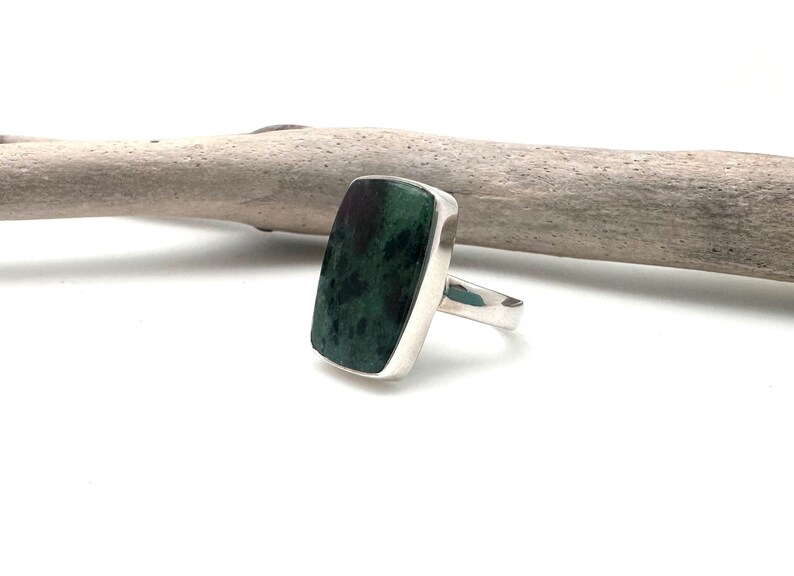 May include: A rectangular ring with a dark green stone and a silver band. The stone has a mottled appearance with dark green and black hues. The ring is set against a white background with a piece of driftwood.