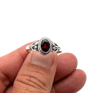 May include: A silver ring with a red gemstone set in a decorative oval bezel. The ring has small silver beads on either side of the gemstone.