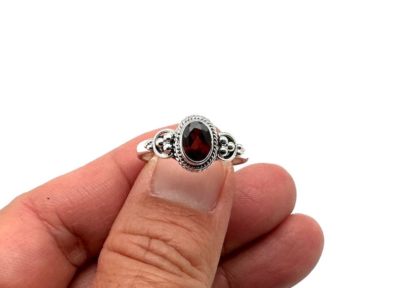 May include: A silver ring with a red gemstone set in a decorative oval bezel. The ring has small silver beads on either side of the gemstone.