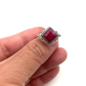 May include: A silver ring with a red gemstone set in a square shape. The ring has a detailed design around the gemstone.