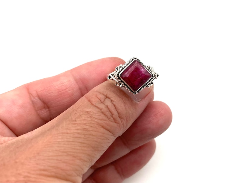 May include: A silver ring with a red gemstone set in a square shape. The ring has a detailed design around the gemstone.