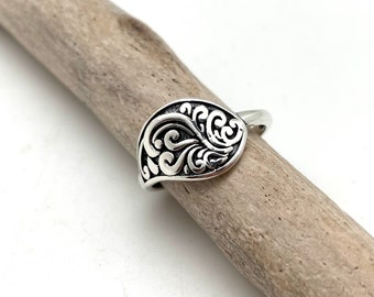 May include: A silver ring featuring a decorative, swirling design. The ring is displayed on a piece of driftwood.