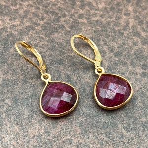 May include: A pair of gold-colored leverback earrings. Each earring features a faceted, teardrop-shaped, deep red gemstone set in a gold bezel. The earrings are displayed against a textured, brown background.