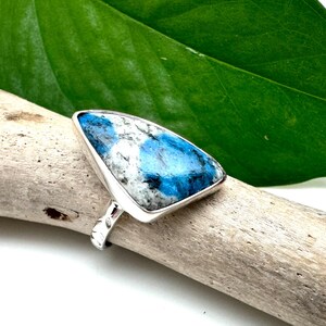 May include: A silver ring with a triangular blue and white gemstone. The ring is set on a piece of driftwood.