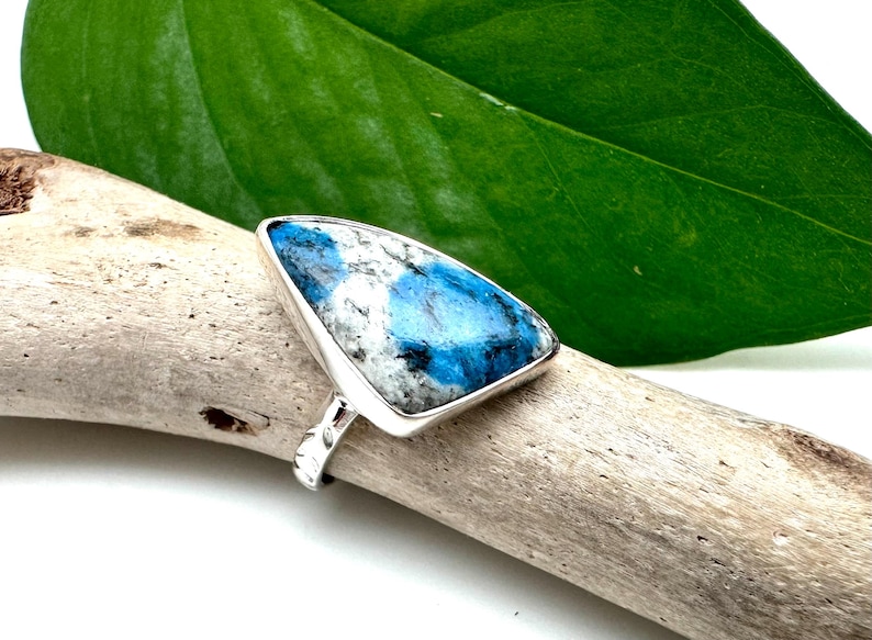 May include: A silver ring with a triangular blue and white gemstone. The ring is set on a piece of driftwood.