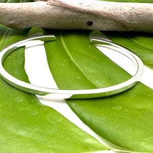 May include: A silver bangle bracelet with a smooth, flat surface. The bracelet is open and has a simple, minimalist design.