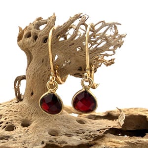 May include: A pair of gold drop earrings featuring faceted, teardrop-shaped deep red garnet stones. Each earring is suspended from a simple gold hook wire, showcasing a polished metal setting that secures the dark red gemstone in a classic drop style.