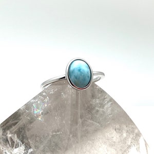 May include: A silver ring featuring a light blue oval-shaped gemstone. The ring is displayed on a white crystal.