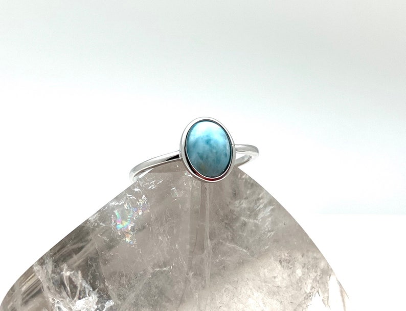 May include: A silver ring featuring a light blue oval-shaped gemstone. The ring is displayed on a white crystal.