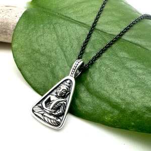 Puede incluir: Un collar con un colgante de plata en forma de triángulo que presenta una imagen detallada de un Buda sentado. El collar tiene una cadena negra.