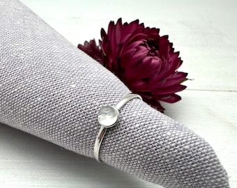 May include: A silver ring with a clear glass stone set in the center. The ring is on a gray fabric background.