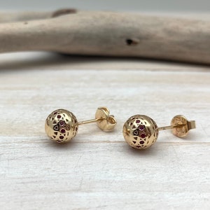 May include: A pair of gold earrings with a round, openwork design and red gemstones. The earrings have a post and butterfly back.