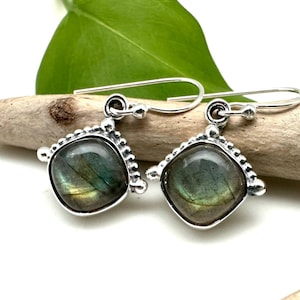 May include: Silver dangle earrings featuring a square-shaped labradorite gemstone. The earrings have a beaded border around the stone.