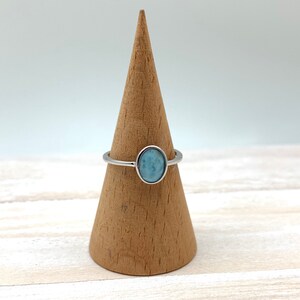 May include: A silver ring with a light blue oval gemstone. The ring is displayed on a wooden ring holder.