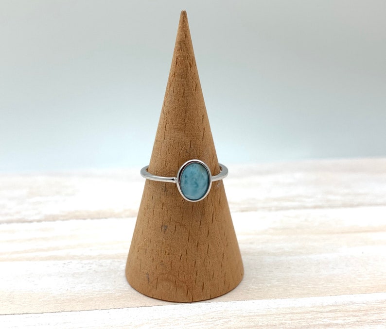 May include: A silver ring with a light blue oval gemstone. The ring is displayed on a wooden ring holder.