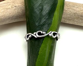 May include: A silver ring with a delicate, swirling vine design. The ring features a repeating pattern of curved lines and small, round accents. The ring is set against a green leaf and a piece of driftwood.