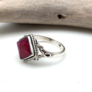 May include: A silver ring with a square red ruby gemstone set in a decorative silver bezel. The ring has a delicate floral design on the band.