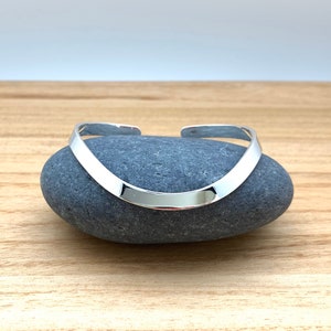 May include: A silver bangle bracelet with a curved, flat design. The bracelet is resting on a gray rock.