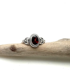 May include: A silver ring with a red gemstone set in a detailed bezel. The ring has a beaded band.