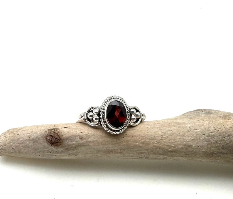 May include: A silver ring with a red gemstone set in a detailed bezel. The ring has a beaded band.