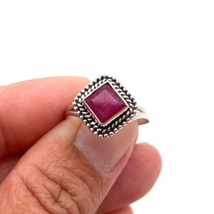 Op de afbeelding: Een zilveren ring met een vierkante robijn-edelsteen gezet in een decoratieve rand. De ring wordt gedragen aan een vinger.