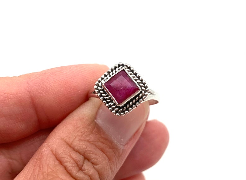 Op de afbeelding: Een zilveren ring met een vierkante robijn-edelsteen gezet in een decoratieve rand. De ring wordt gedragen aan een vinger.